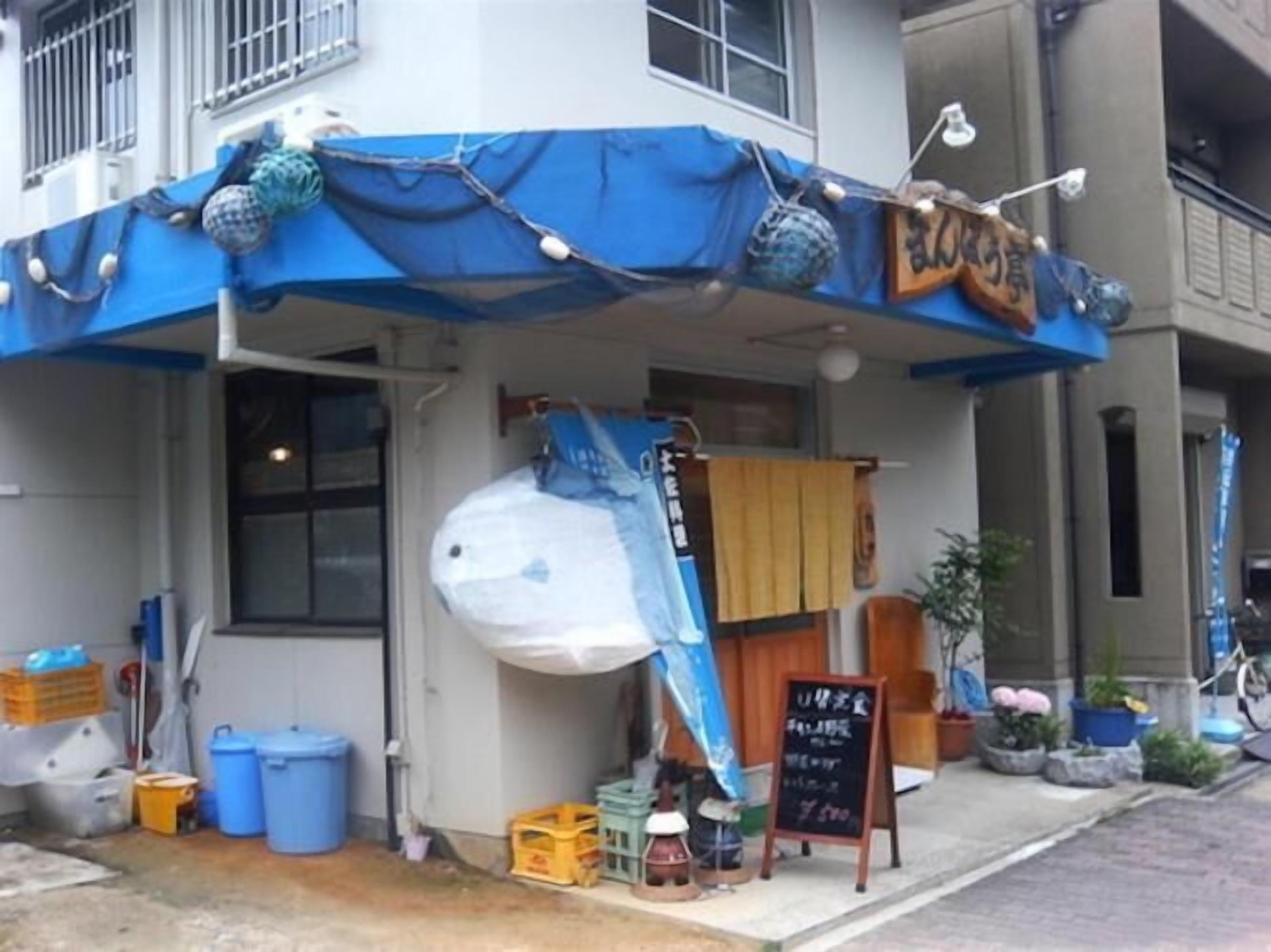 A storefront with a blue awning, a signboard, and various items outside including a large white object, blue barrels, and a yellow crate.