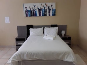 Ett hotellrum med en stor säng, två sängbord, ett litet bord, en tavla på väggen och en vit handduk på sängen.