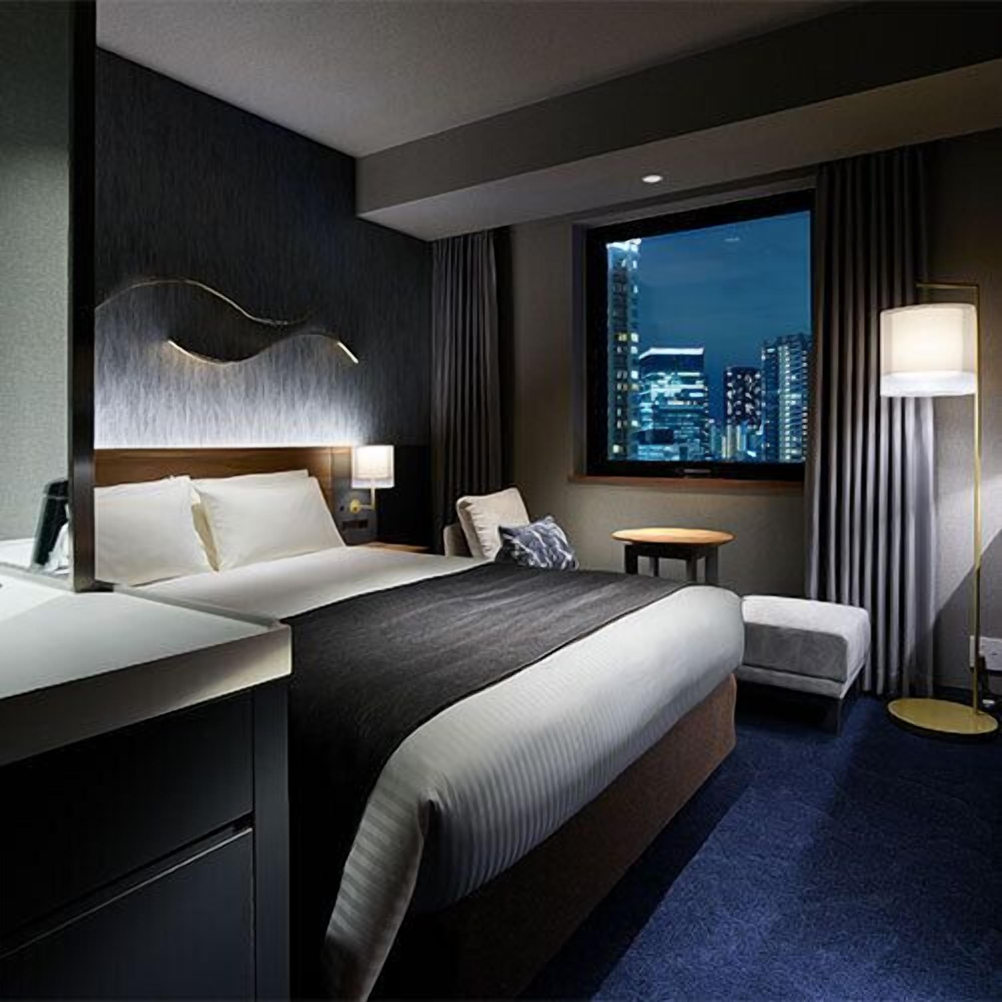 A modern hotel room with a large bed, a desk, a chair, a small table, and a view of a cityscape at night.