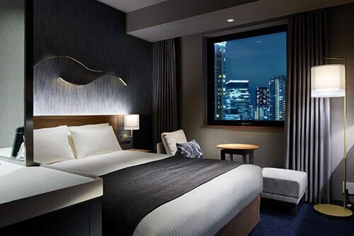A modern hotel room with a large bed, a desk, a chair, a small table, and a view of a cityscape at night.