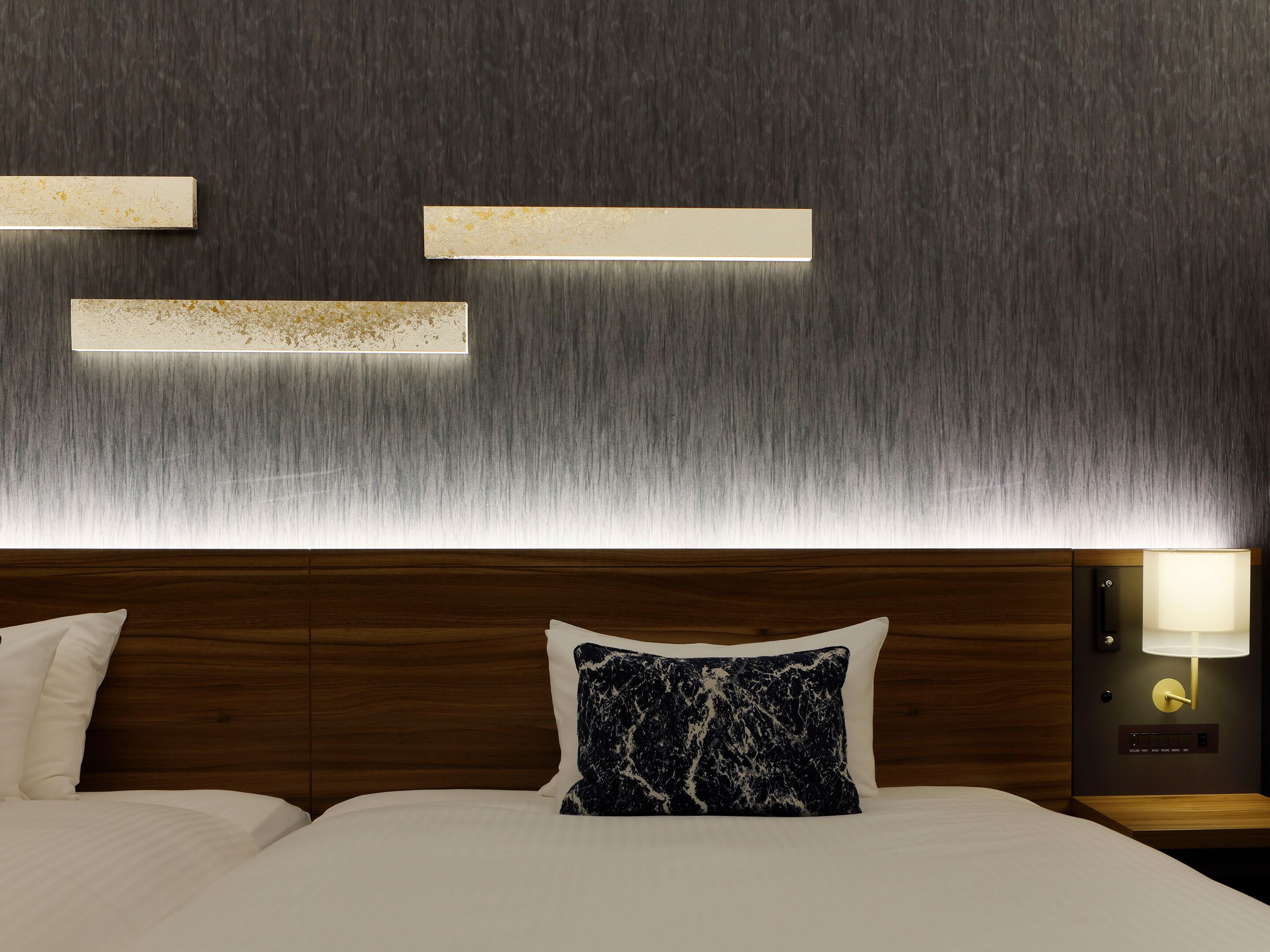 A hotel room with a wooden headboard, a bed with white bedding, a marble-patterned pillow, and wall-mounted lighting.