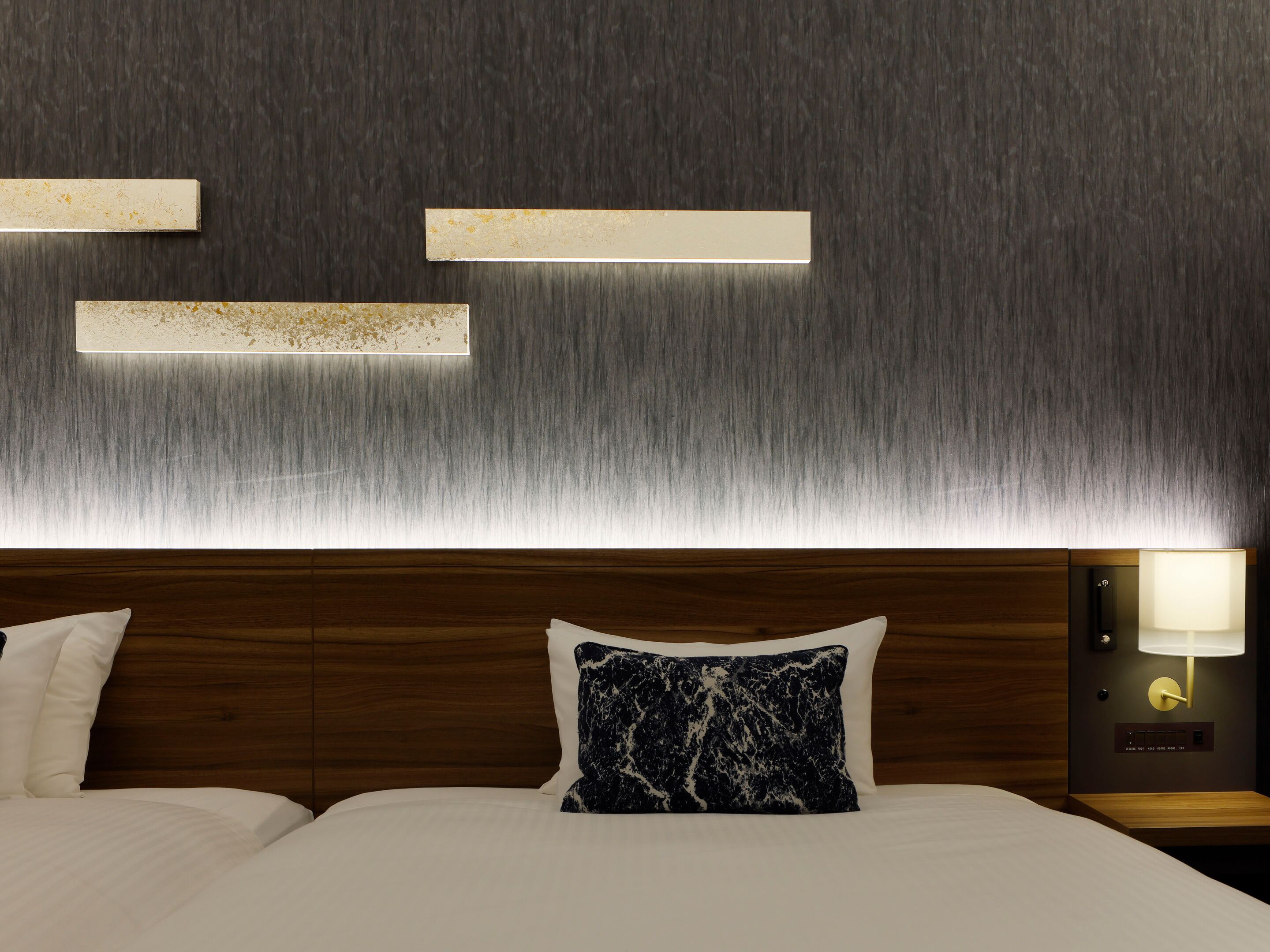 A hotel room with a wooden headboard, a marble-patterned pillow, a nightstand with a lamp, and wall-mounted light fixtures.