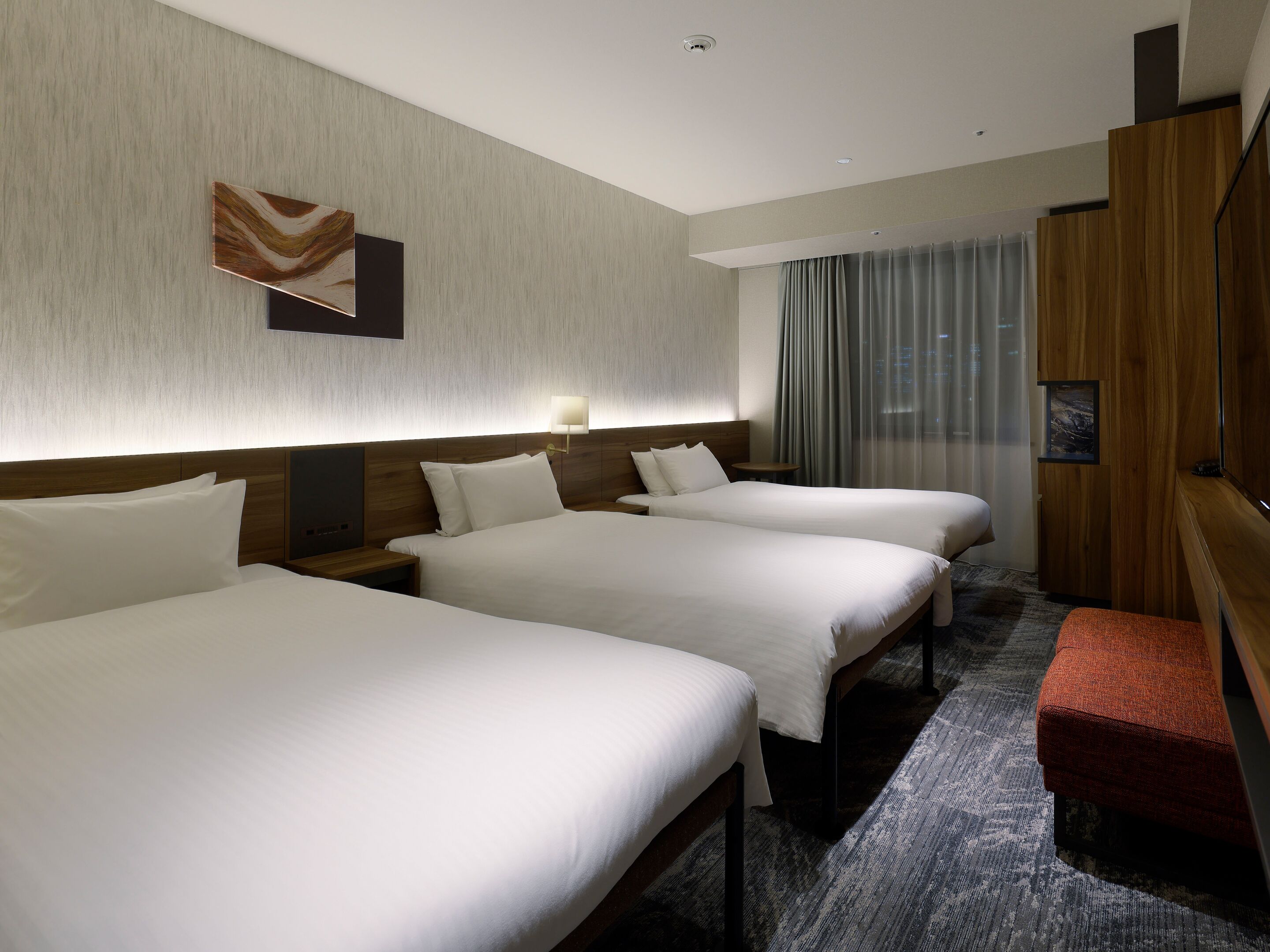 A hotel room with three beds, a wooden headboard, a small table, and a bench.