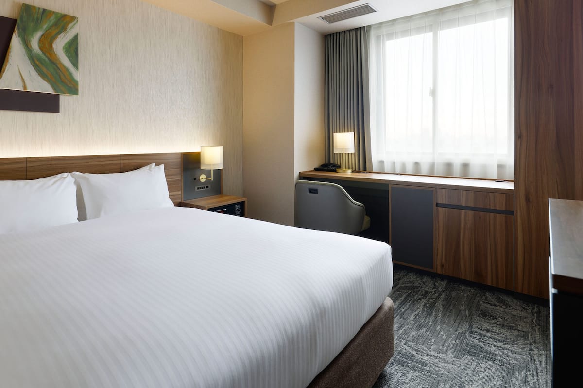Ein Hotelzimmer mit einem großen Bett, einem Schreibtisch mit Lampe, einem Sessel und einem Fenster mit Vorhängen.