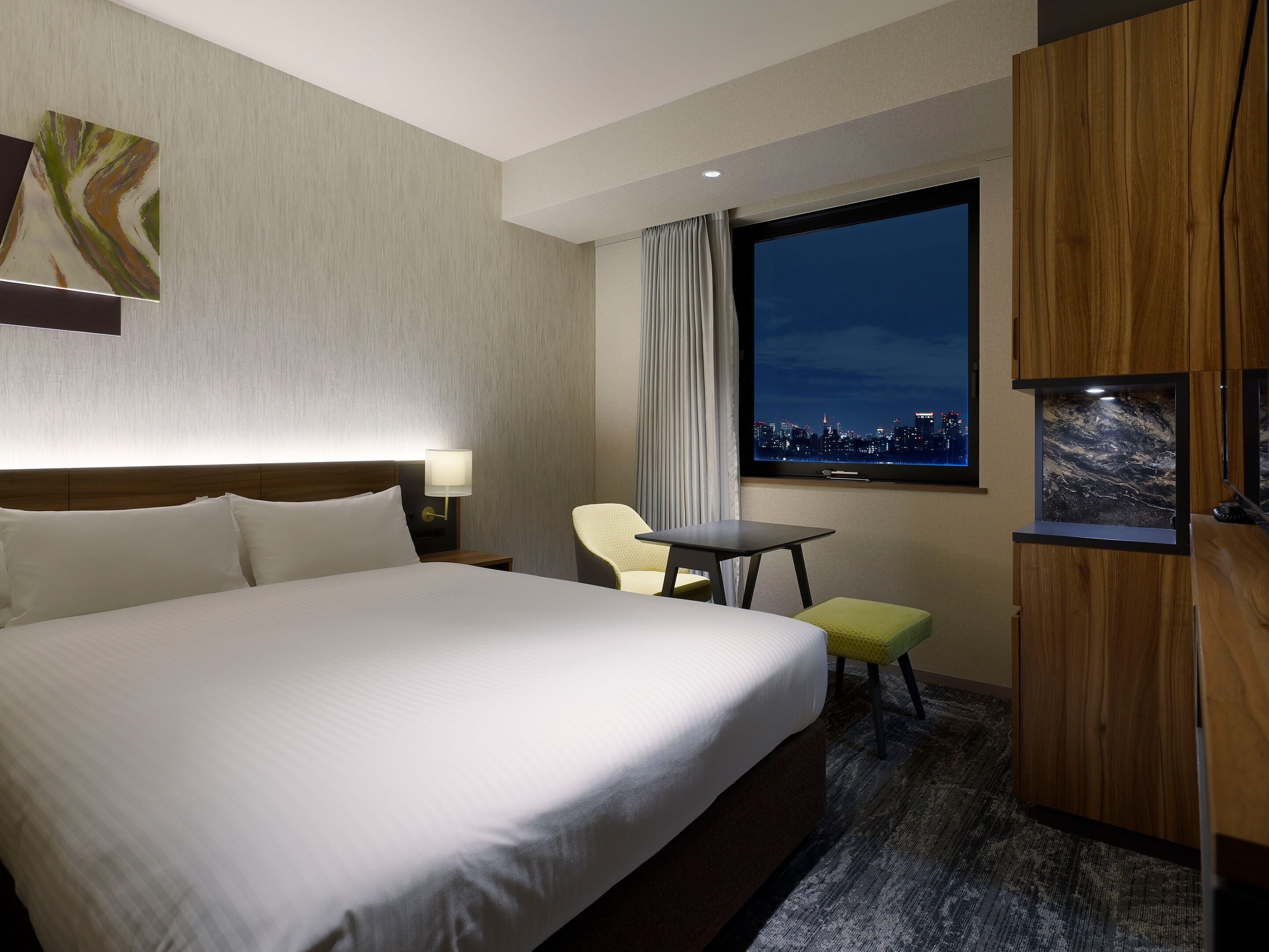 A hotel room with a large bed, a small table, a chair, a TV, and a city view.