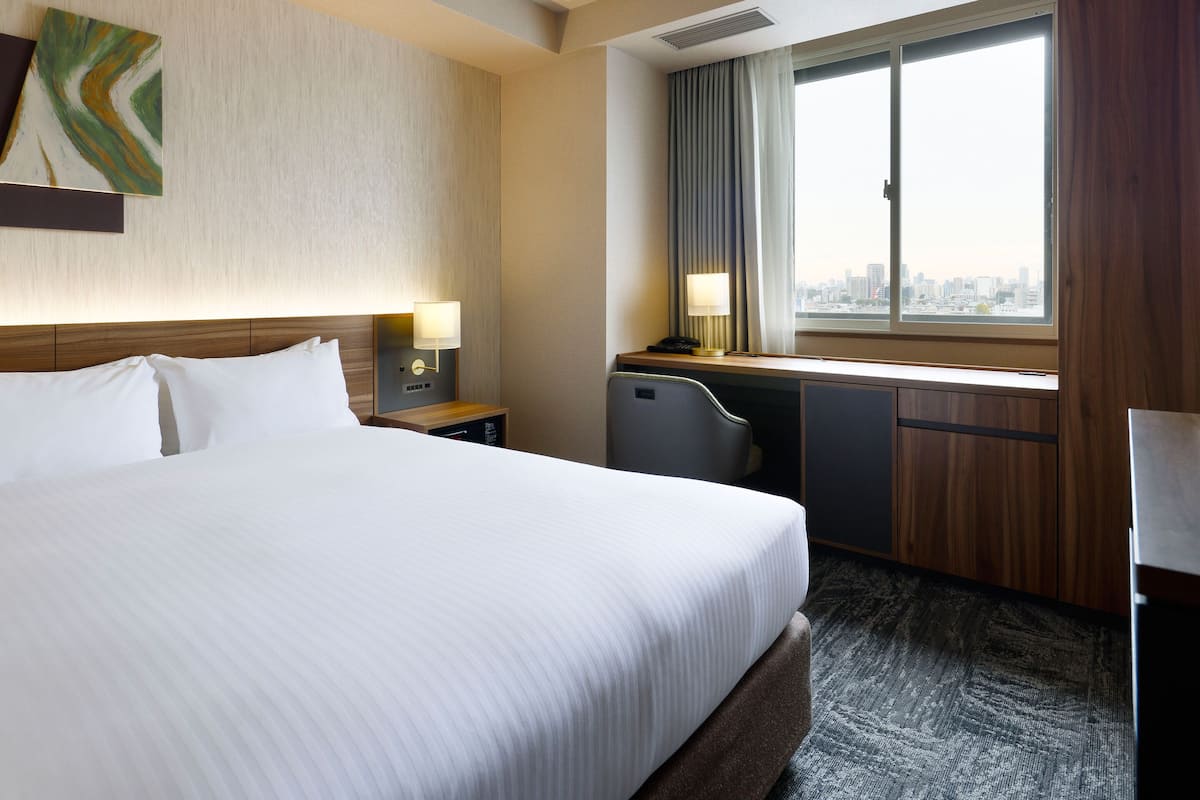 Ein Hotelzimmer mit Bett, Schreibtisch, Stuhl, Lampe und einem Fenster mit Blick auf die Stadt.