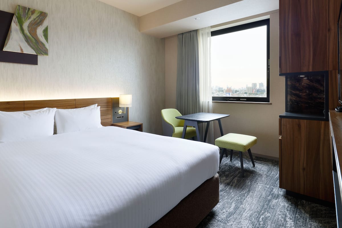 Ein Hotelzimmer mit einem großen Bett, einem kleinen Tisch, zwei Stühlen, einem Fernseher und einem Fenster mit Blick auf die Stadt.