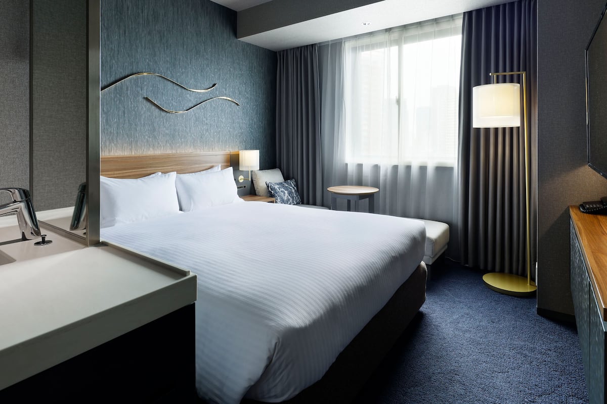Ein Hotelzimmer mit Bett, Nachttisch, Lampe und Blick ins Freie durch ein Fenster.