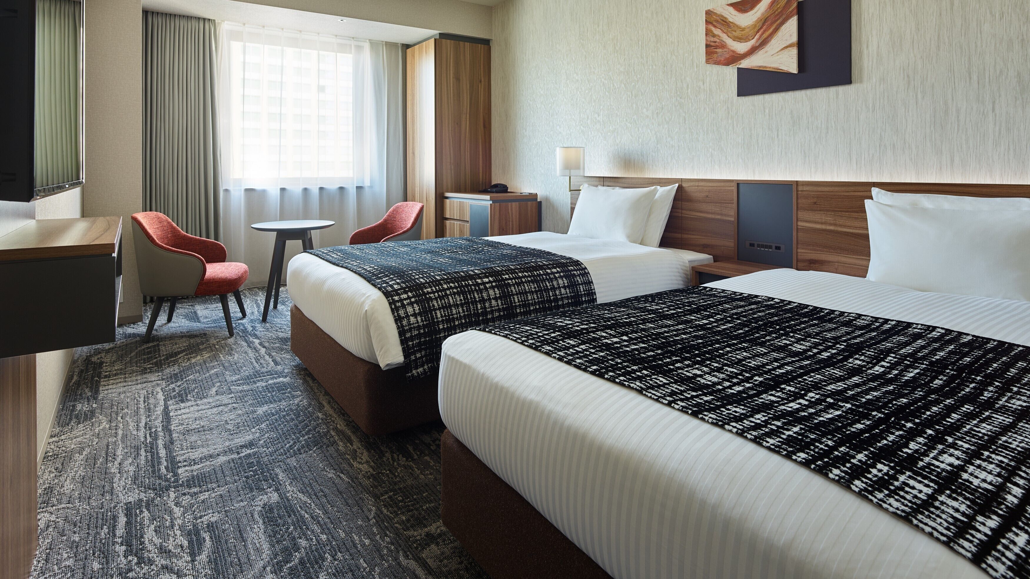 A hotel room with two beds, a desk, a chair, a small table, and a lamp.