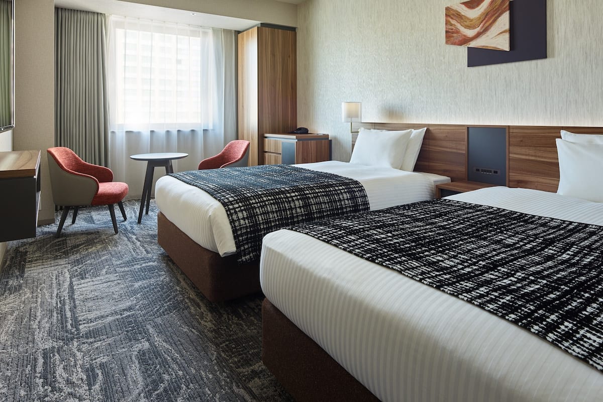 A hotel room with two beds, a desk, a chair, a small table, and a lamp.