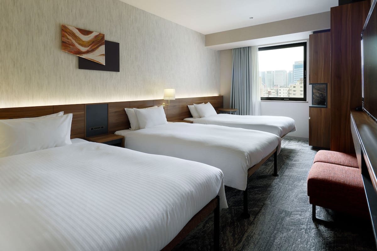 Ein Hotelzimmer mit drei Betten, einem Holzkopfende, Blick auf die Stadt, einem kleinen Tisch und einer roten Bank.