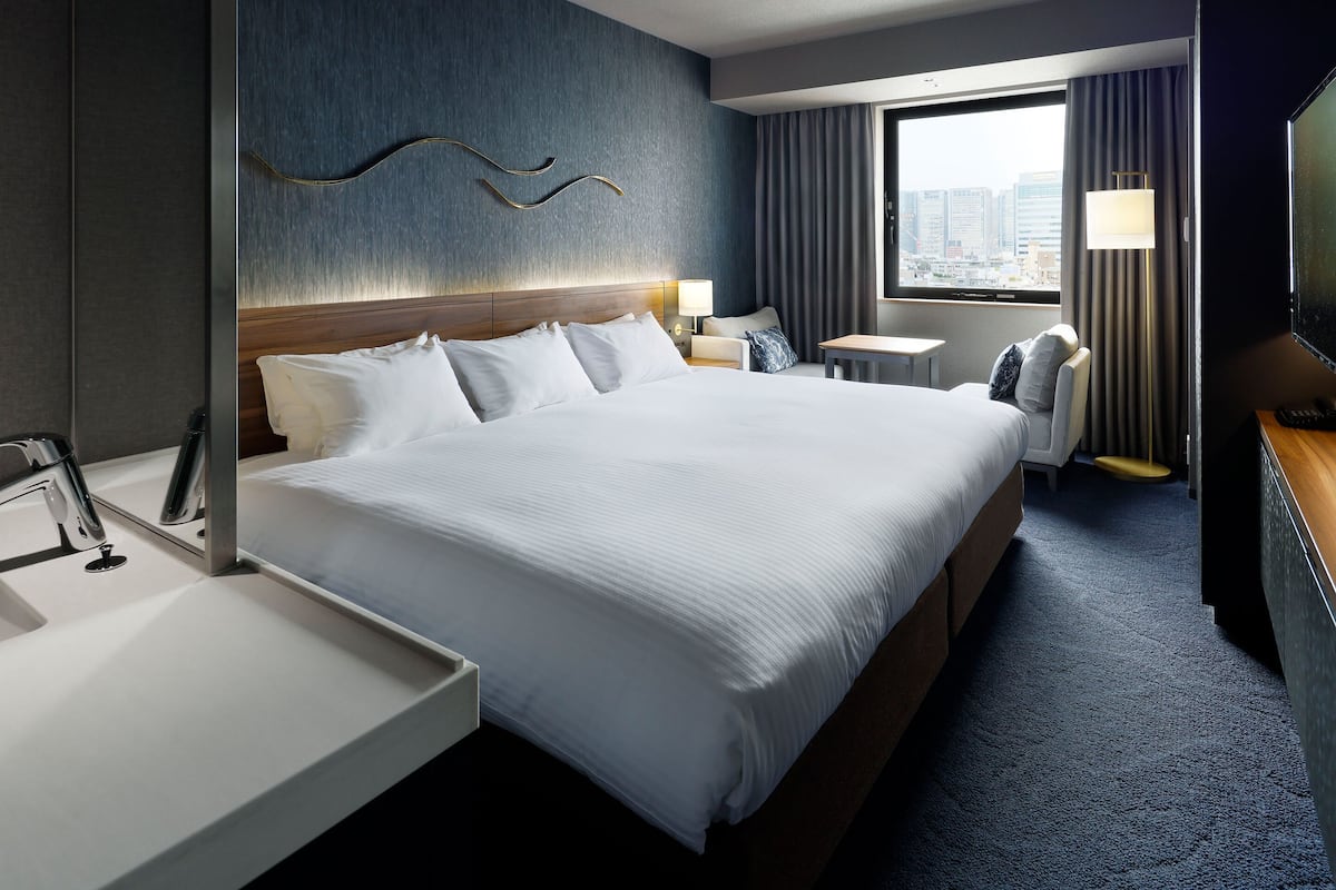 Ein Hotelzimmer mit einem großen Bett, einem Nachttisch, einem Schreibtisch, einem Sessel, einem Fernseher und einem Fenster mit Vorhängen.