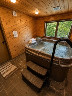 A wooden cabin interior with a hot tub, a window with a view of greenery, and a wooden floor.