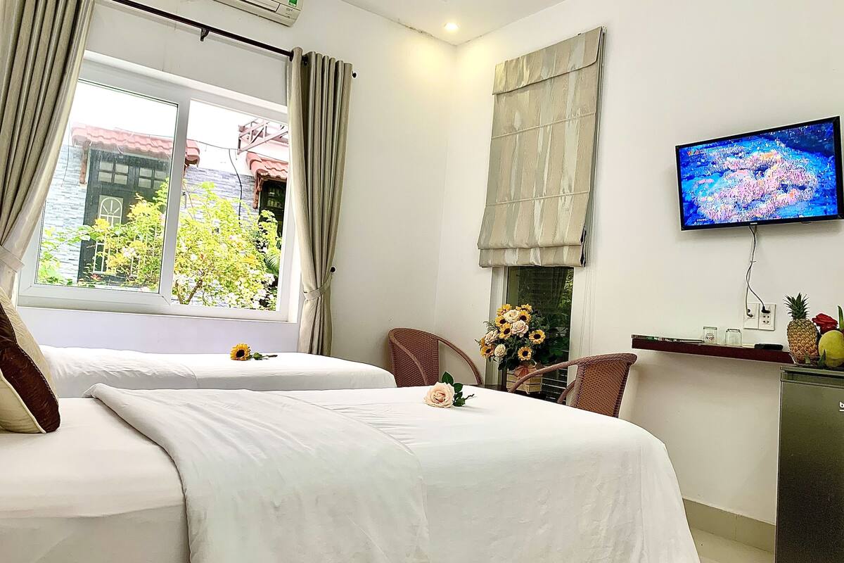 Habitación de hotel con dos camas, televisor, escritorio con piña y flores, y ventana con cortinas.
