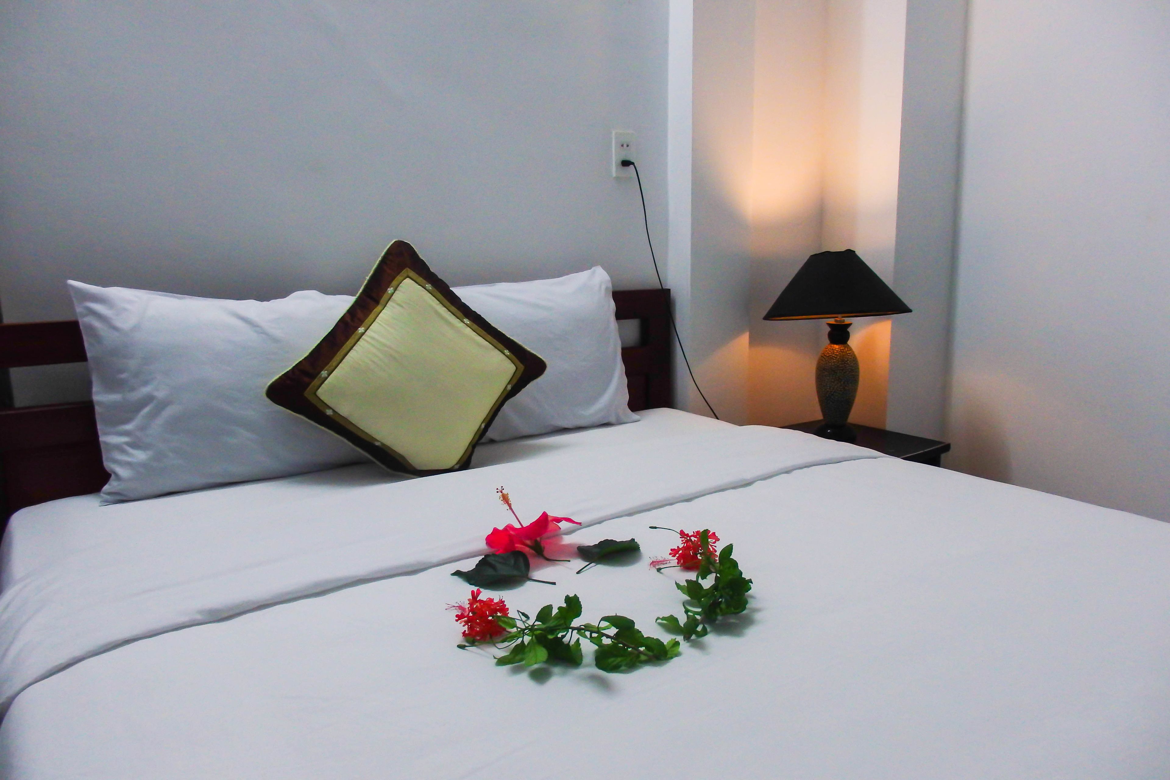 Una cama bien hecha con sábanas y almohadas blancas, un arreglo floral decorativo sobre la cama y una lámpara de noche.