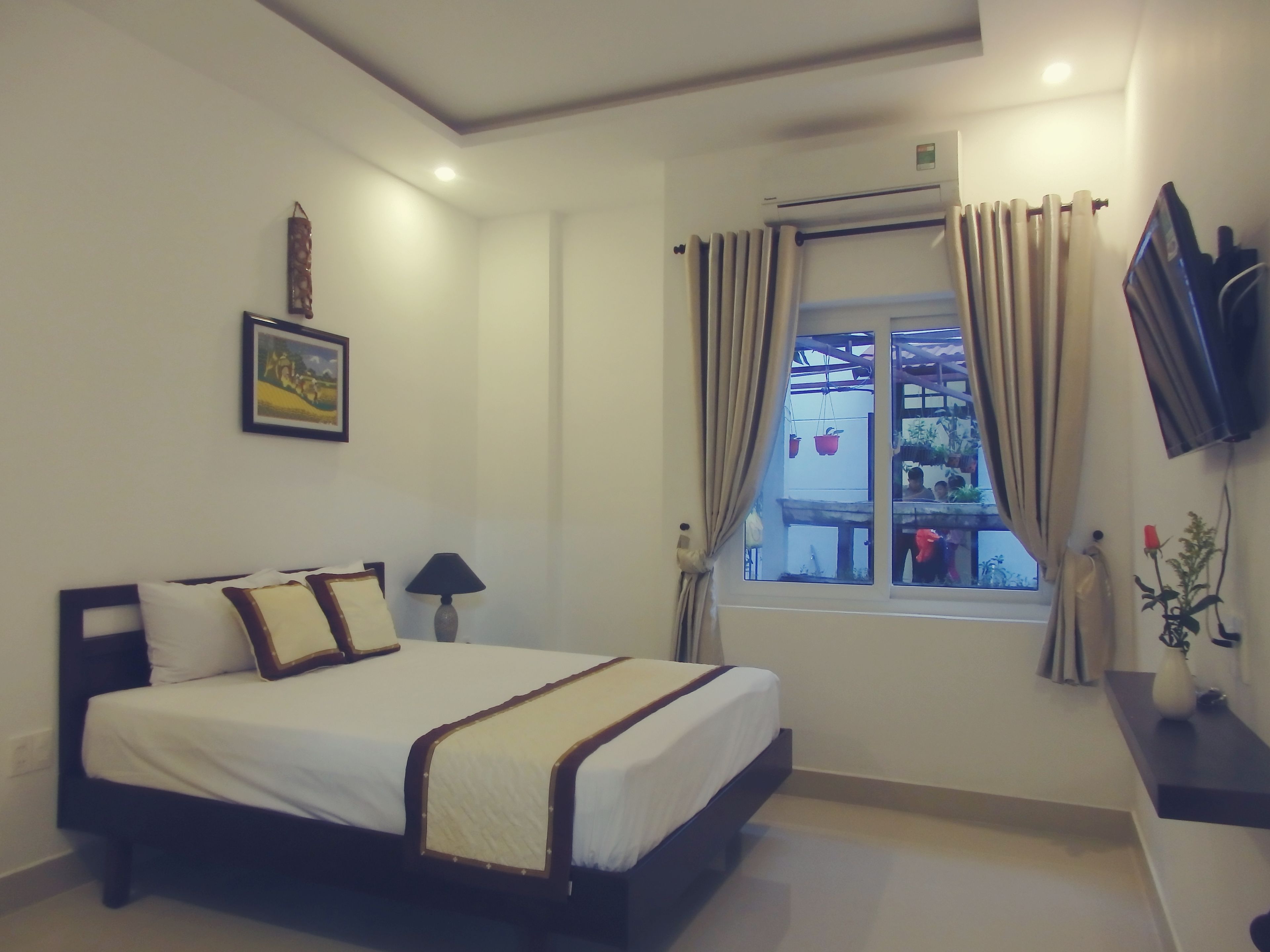 A bedroom with a bed, a TV mounted on the wall, a window with curtains, and a small table with a vase.