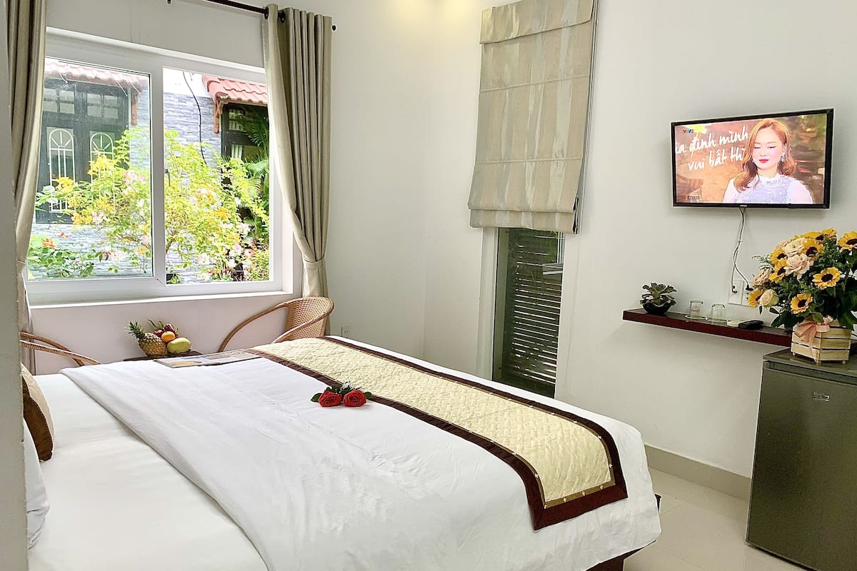 Una cama bien tendida con edredón blanco y color beige, un televisor montado en la pared y una ventana con cortinas.