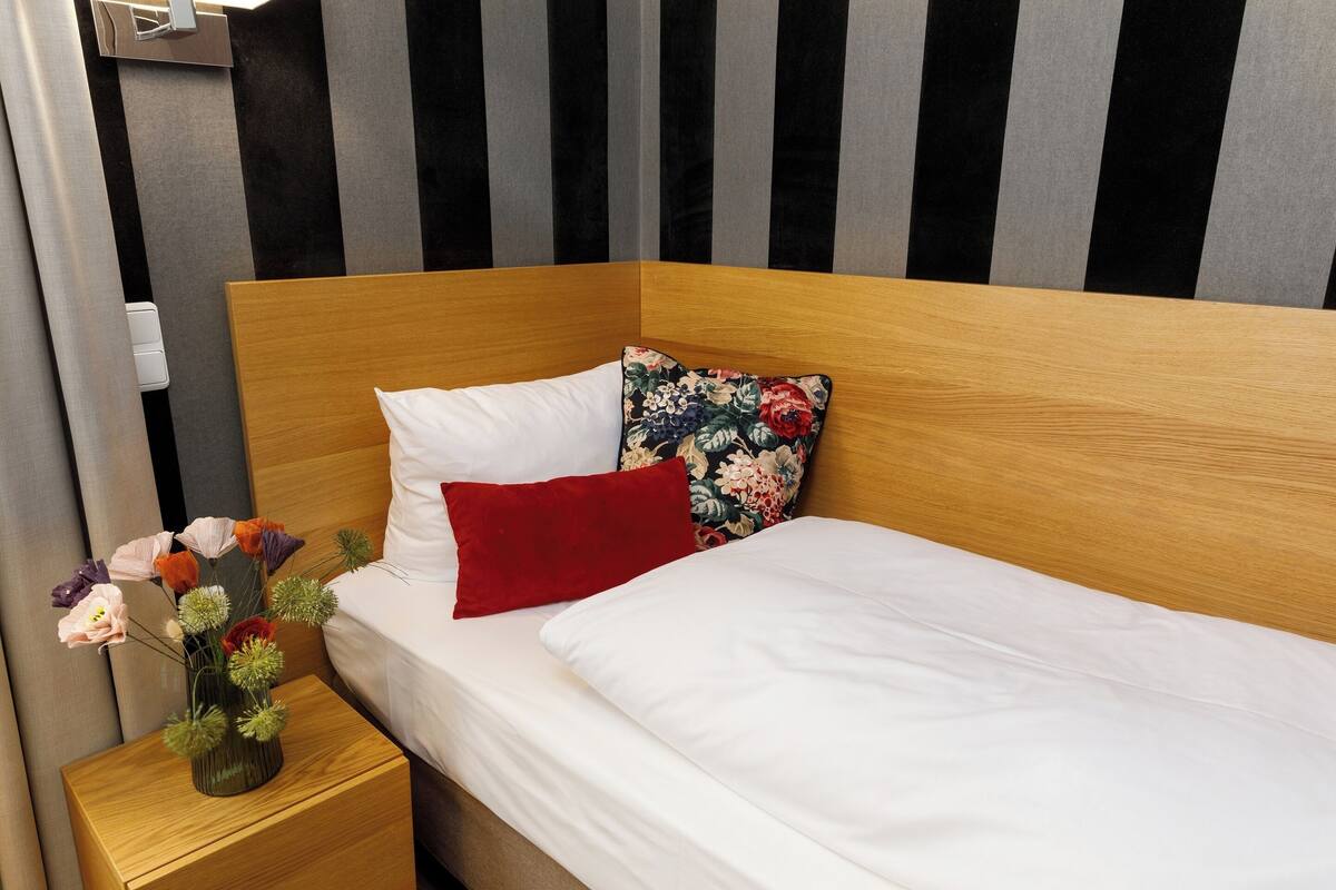 A neatly made bed with white linens, a wooden headboard, and a nightstand with a vase of flowers.