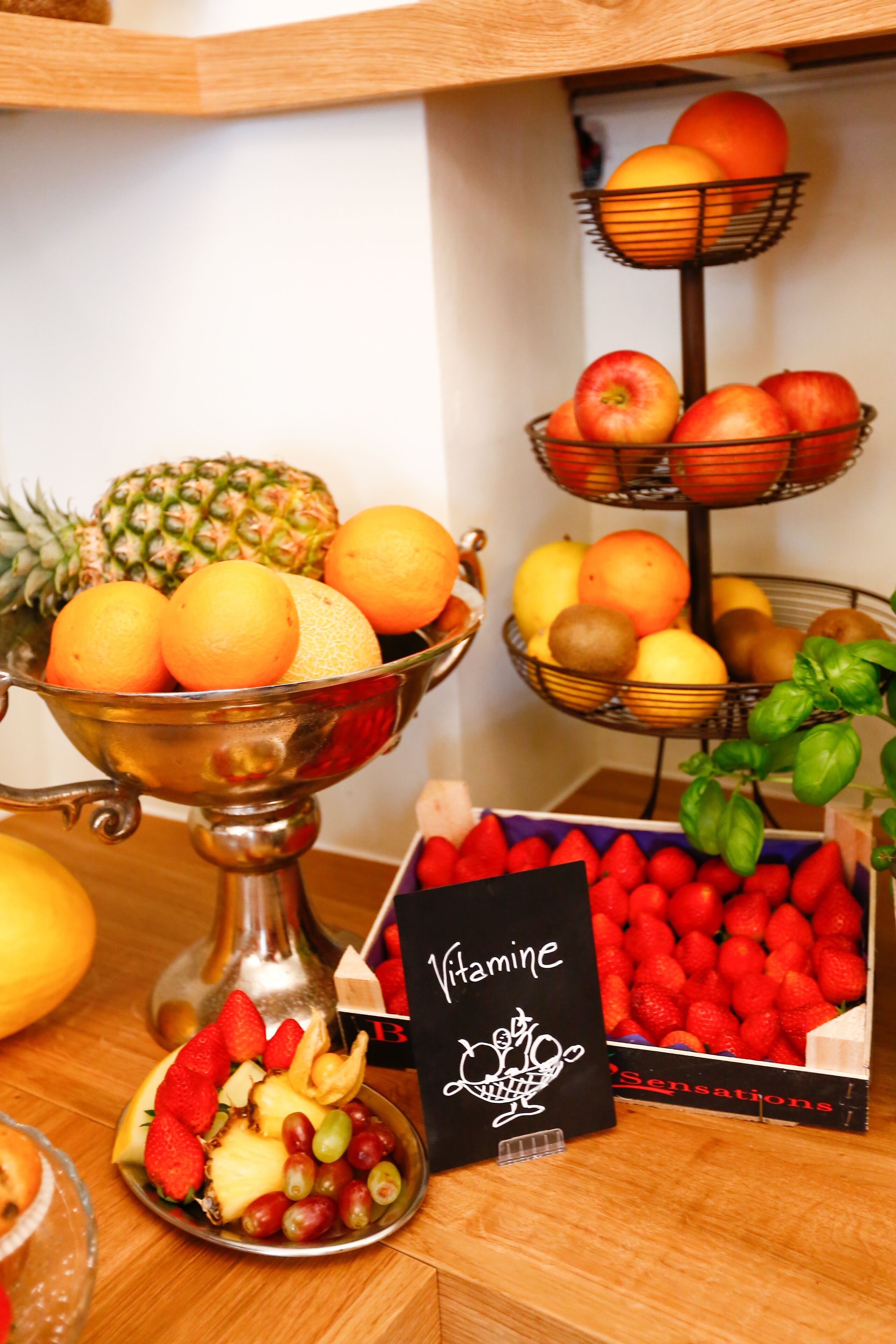 Eine Obstauslage mit verschiedenen Früchten in Schalen und Körben, ein Schild mit der Aufschrift "Vitamin B" und eine Kiste Erdbeeren.