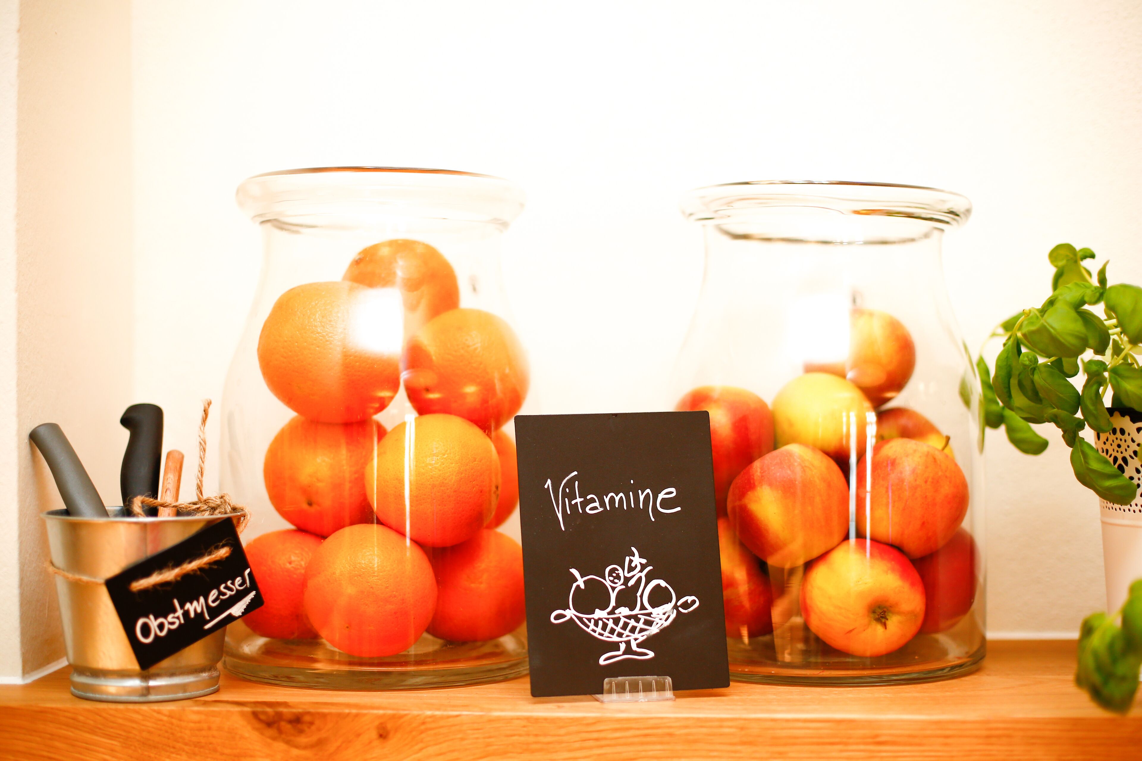 Two glass jars filled with oranges and apples, a metal container with a label, and a card with the word "Vitamine".