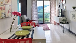 A dining area with a table set for two, red chairs, and a vase with flowers. There is a large window with curtains, a small table with a plant, and a wall with framed pictures and a sign.