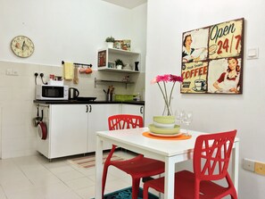Una pequeña cocina con armarios blancos, un microondas y una cafetera. Hay una mesa de comedor con sillas rojas y un jarrón con flores. Un anuncio de café antiguo está en la pared.