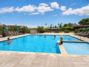 A swimming pool with lounge chairs and umbrellas, surrounded by palm trees and buildings.
