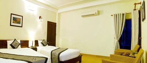 A hotel room with two beds, a sitting area with a chair, a wall-mounted air conditioner, and a framed picture on the wall.