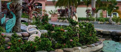 Un jardín ajardinado con un estanque, esculturas decorativas y un edificio con tejado de tejas al fondo.