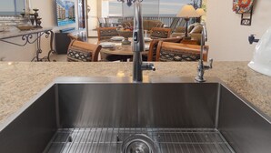 Una cocina con un lavamanos de acero inoxidable, un comedor con mesa y sillas, y vista al océano por la ventana.