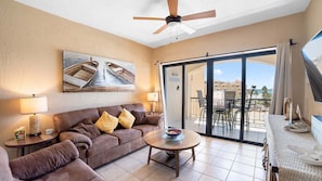 A living room with a sofa, a coffee table, a ceiling fan, and a large window with a view of buildings and a beach.