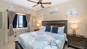 A bedroom with a bed, a ceiling fan, artwork on the walls, and a window with curtains.