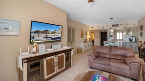 A living room with a brown sofa, a television displaying a beach scene, a dining area with a kitchen in the background, and a wall decorated with fish and a welcome sign.