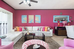A living room with a white sofa, colorful cushions, a glass coffee table, and a pink chair. There are two abstract paintings on the wall, a ceiling fan, and a mirror above a wooden cabinet.
