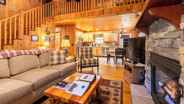 Ein gemütliches Blockhaus-Interieur mit einer Holztreppe, einem Wohnzimmer mit Sofa und Couchtisch, einem Essbereich und einem Kamin aus Stein.