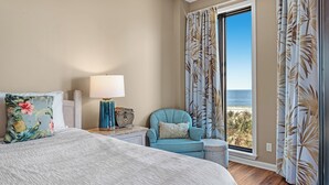 Una habitación de hotel con cama, sillón, mesita de noche, lámpara y vista a la playa.