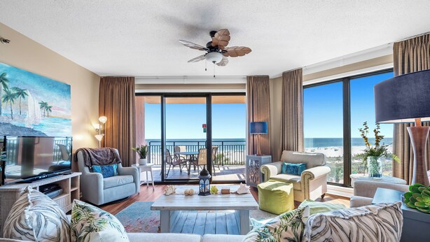 Un salón amplio con un televisor de pantalla plana grande, asientos cómodos, un ventilador de techo y un balcón con vista a la playa.
