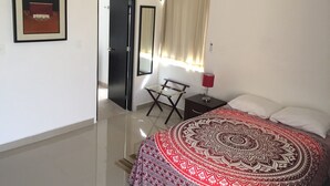 Quarto com cama, ventilador de teto, mesa de cabeceira com luminária, quadro na parede e porta para outro cômodo.