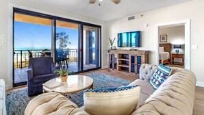 Ein Wohnzimmer mit einer Couch, einem Sessel, einem Couchtisch und einem Fernseher. Ein Balkon bietet Blick auf den Ozean.