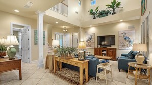 A spacious living room with a sofa, a coffee table, and a TV. There are potted plants and artwork on the walls.