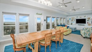 Un salón espacioso con una mesa de comedor de madera, asientos cómodos, un televisor de pantalla plana grande y vistas a la playa a través de puertas corredizas de vidrio.