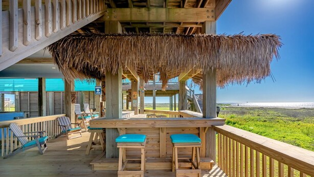 Una terraza de madera con techo de paja, dos taburetes de bar y vista al océano.