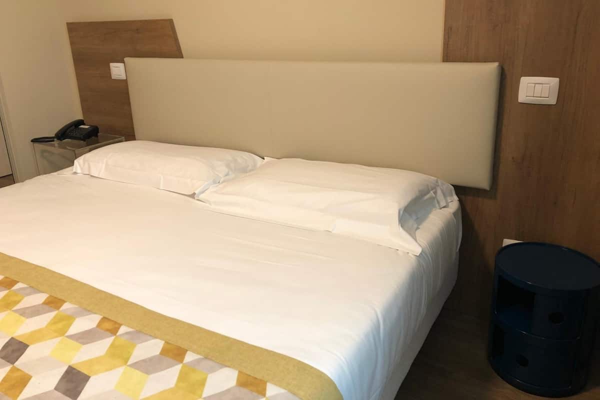 A hotel room with a large bed, a wooden headboard, a bedside table with a phone, and a trash bin.