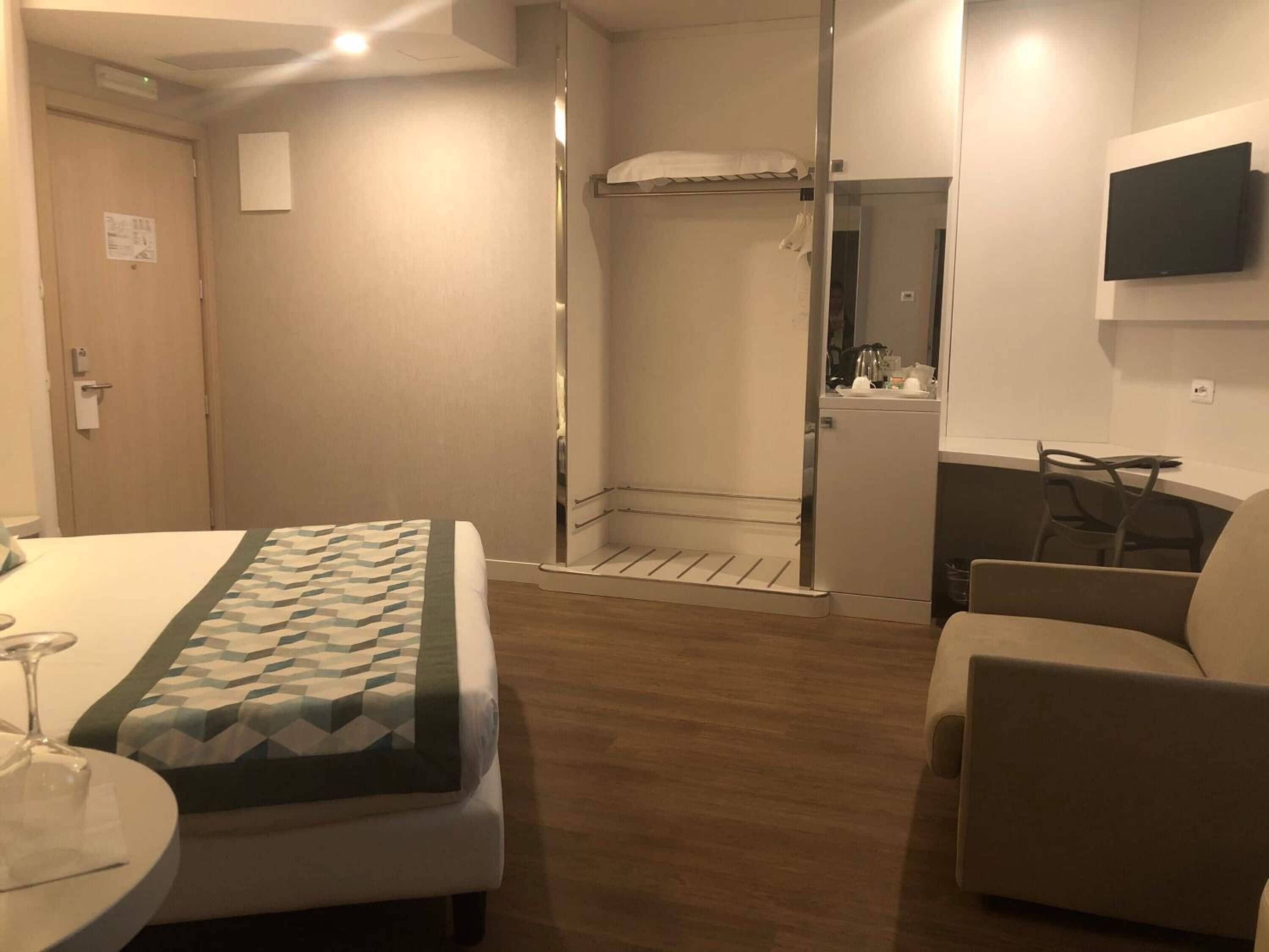 A hotel room with a bed, a small table with a glass, a chair, a sofa, a TV mounted on the wall, and a shower area.