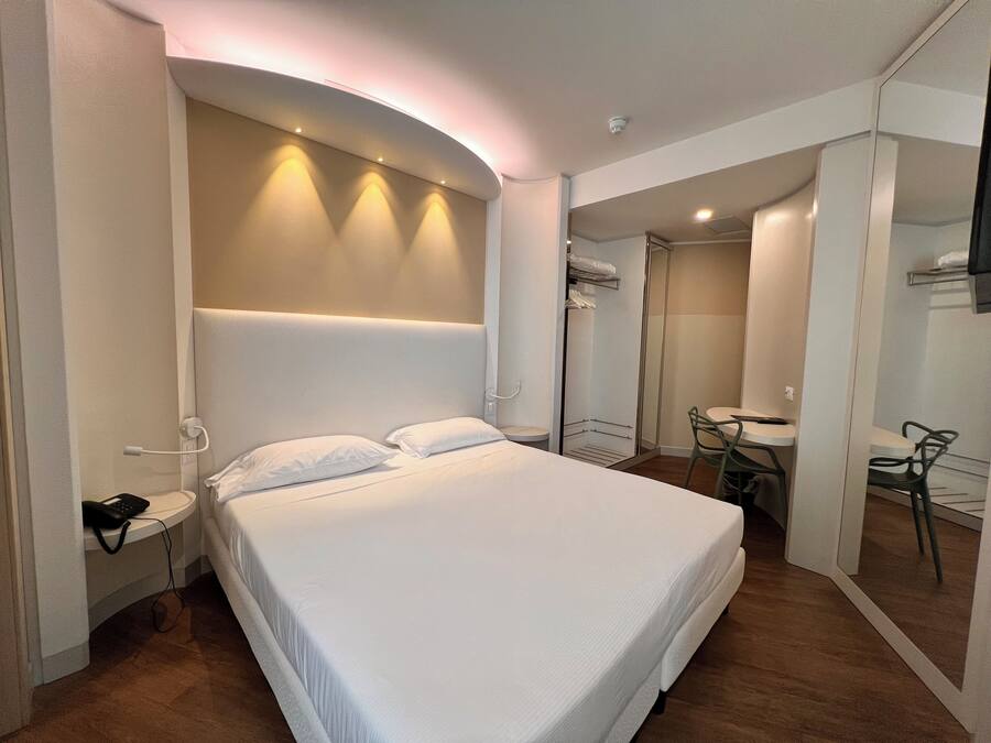 A modern hotel room with a large bed, bedside tables, a small desk, and a bathroom visible through a glass partition.
