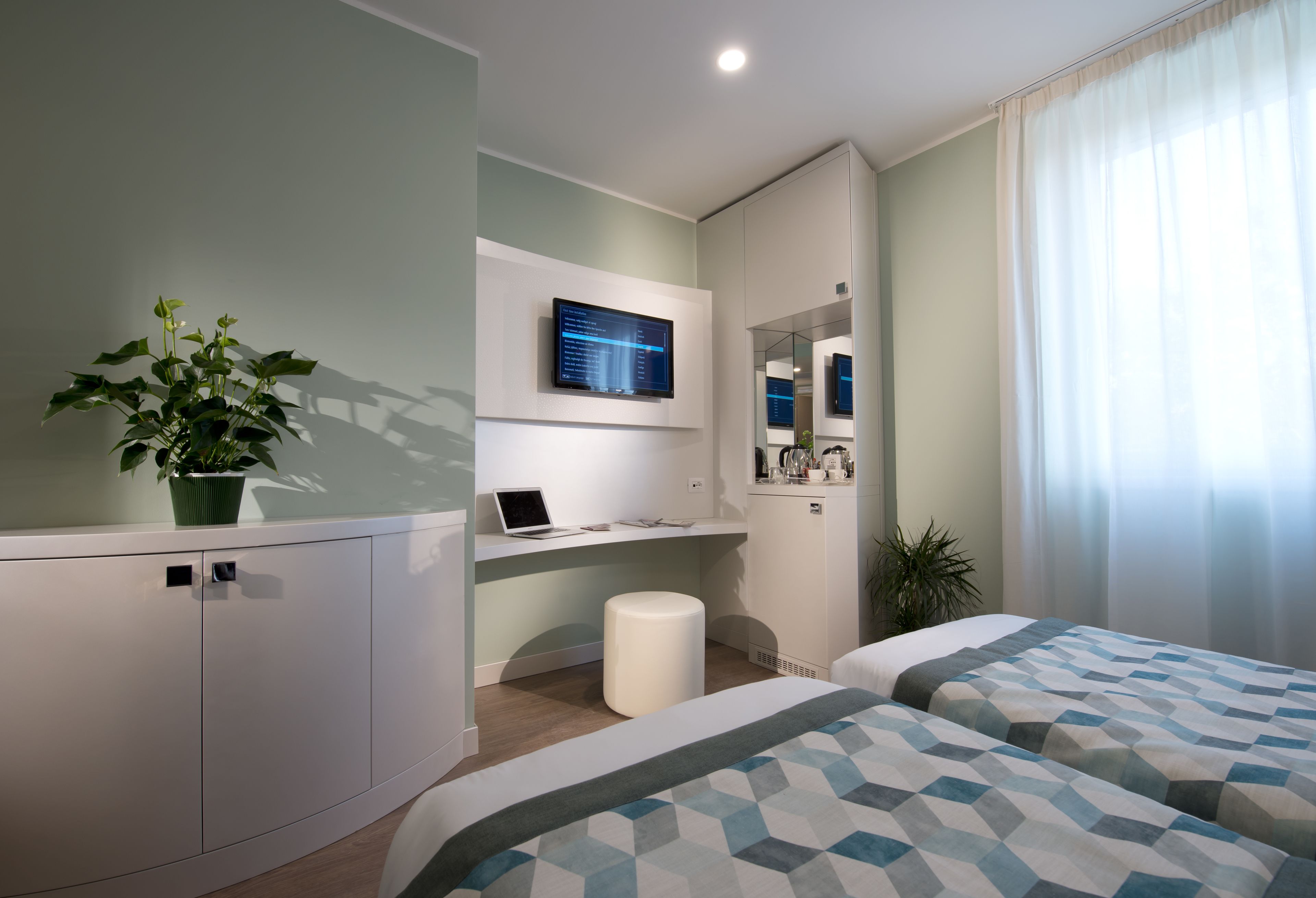 Ett modernt hotellrum med en säng, ett skrivbord med en bärbar dator, en platt-TV, en krukväxt och en spegel.