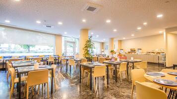 A spacious dining area with multiple tables and chairs, large windows, and a kitchen in the background.