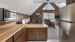 A modern kitchen with wooden cabinetry, a stainless steel refrigerator, and a dining area with a large window. - A Cozy Vacation Home with a Sauna & Beautiful Nature View (Ironwood)