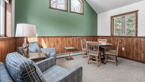 A room with a green wall, wooden paneling, a plaid sofa, a wooden table with chairs, and two windows. - A Cozy Vacation Home with a Sauna & Beautiful Nature View (Ironwood)