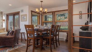 A dining area with a wooden table and chairs, a chandelier, and large windows with a view of the outdoors. - JHRL - Nez Perce D2, Recently updated 3-bedroom Mountain Decor - 1 mile from Grand Teton NP (Teton Village)